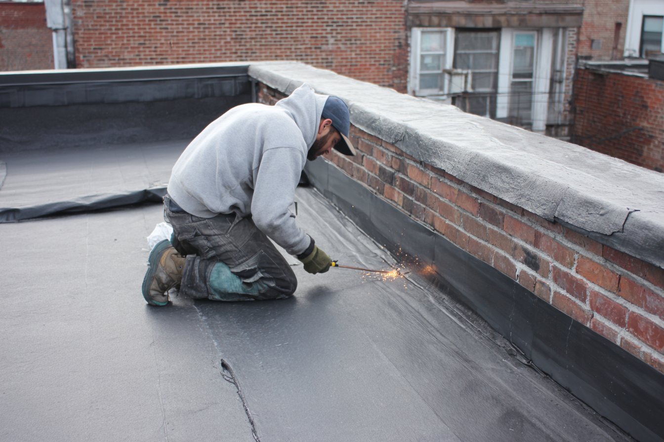 Flat Roofers NY roofer working on the flat modified bitumen roof of an attached Astoria rowhouse, parapet wall and neighbouring roofs in view