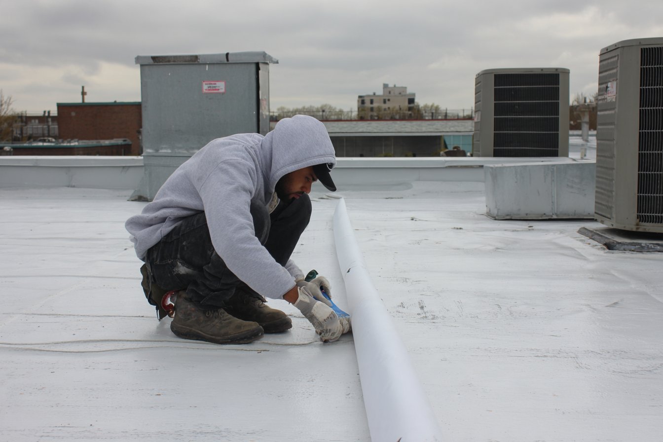 Roofer resealing a seam on the white membrane roof of a Queens commercial building, rooftop HVAC units behind him