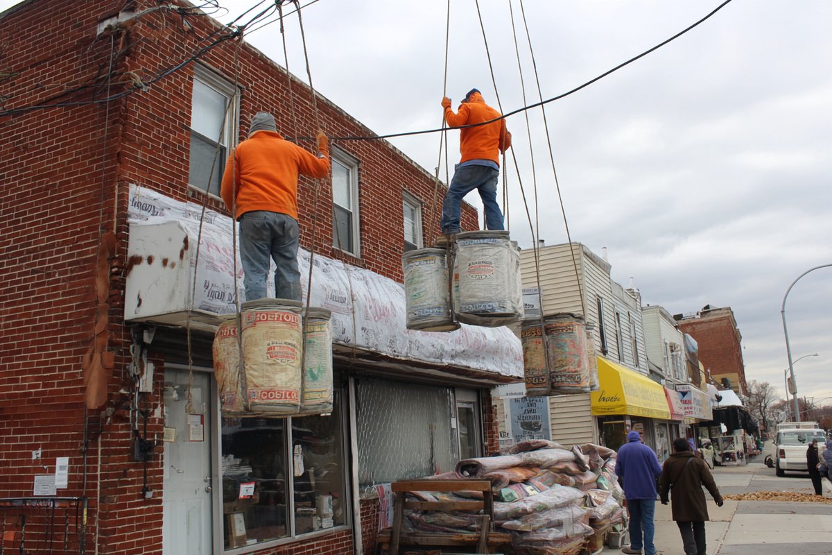 Roofing materials hoisted up the outside of an open Queens storefront building while customers come and go below