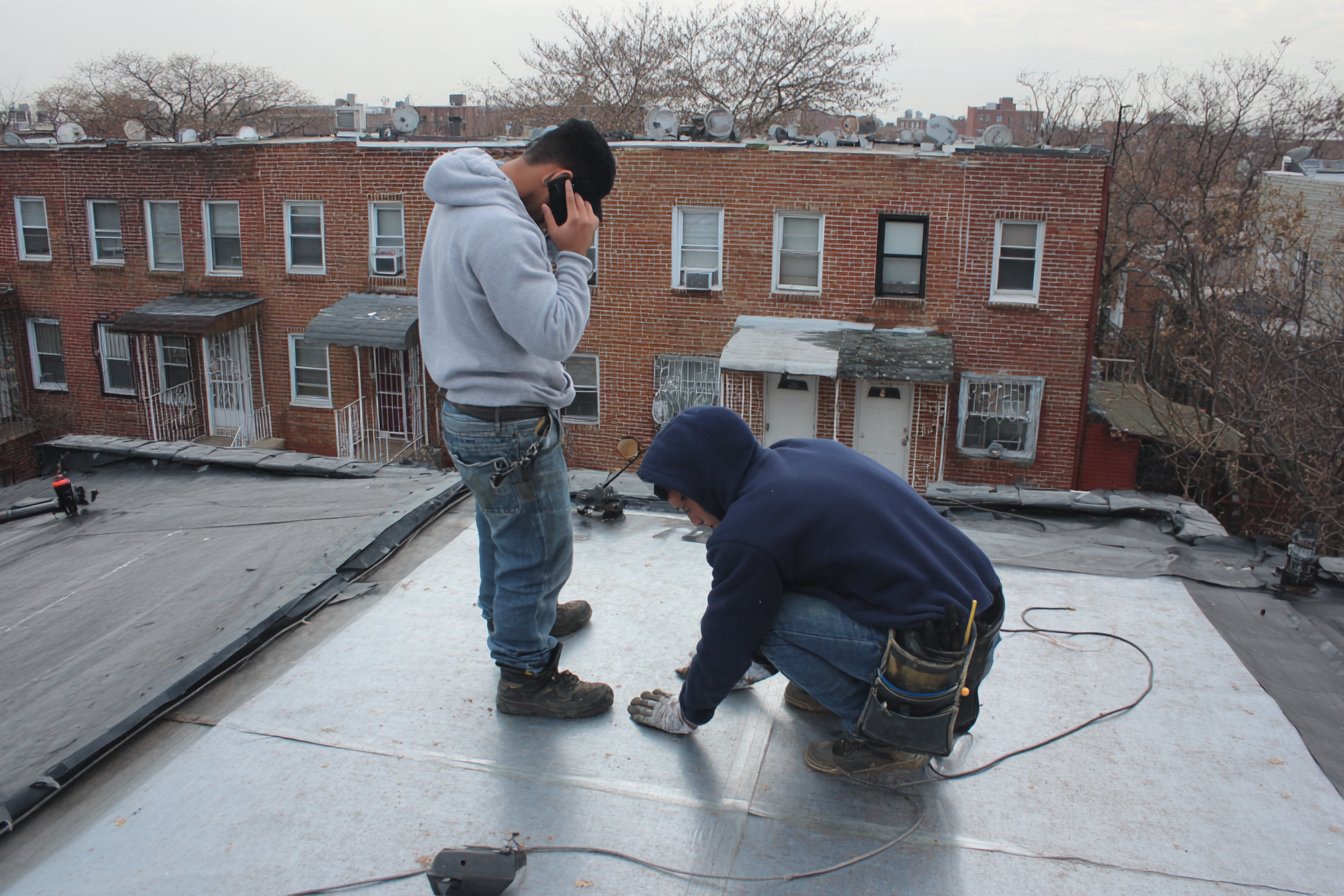 Flat Roofers NY roofer on a Queens flat roof taking a customer's call beside the roof hatch