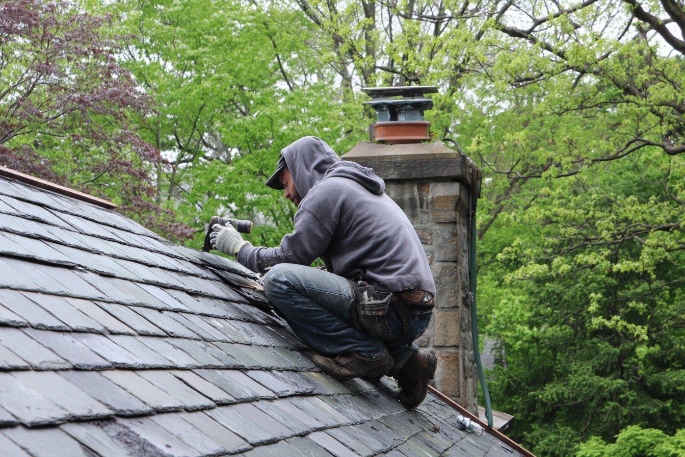 Flat Roofers NY roofer resetting slate on the steep front pitch of a Tudor house in Douglaston, Queens, with mature street trees behind