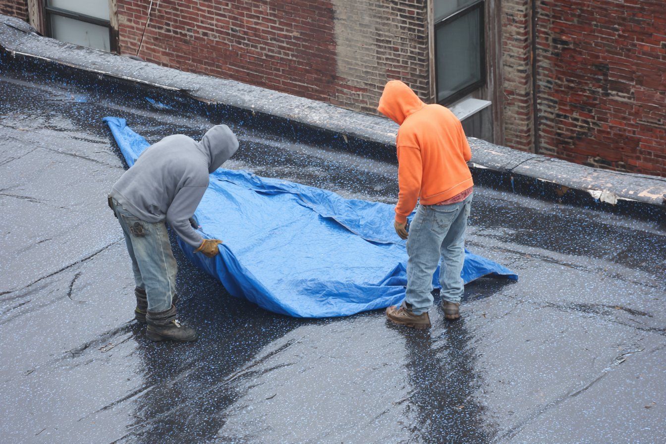 Flat Roofers NY crew laying an emergency tarp over a leaking flat roof on a Queens brownstone during rain