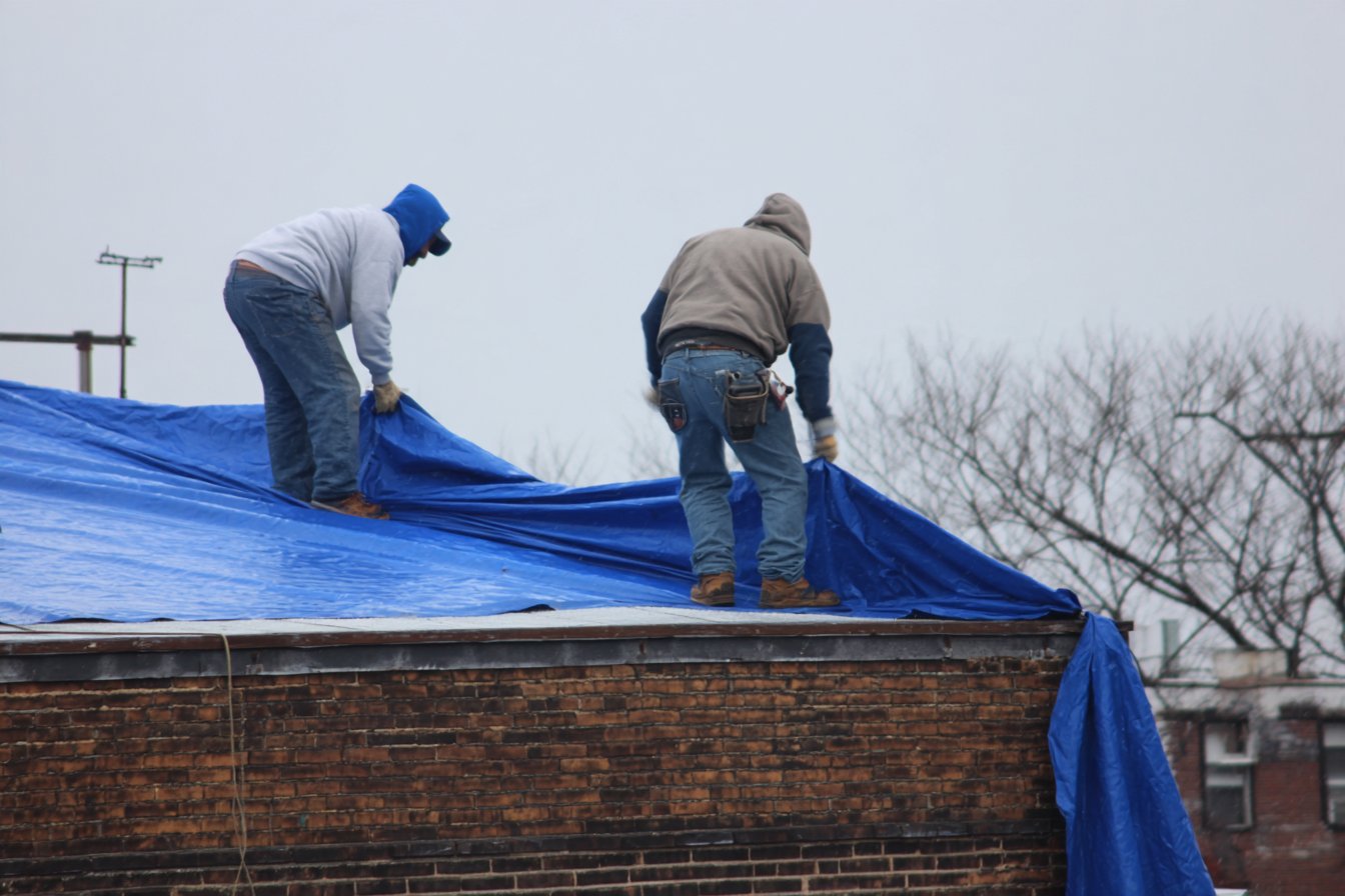 Flat Roofers NY crew stretching a weighted emergency tarp over a storm-torn flat roof on a Queens building