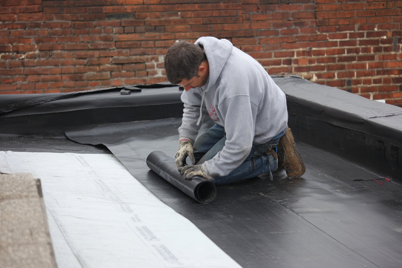 Roofer rolling out a new black EPDM rubber membrane over polyiso insulation on a Queens row house flat roof