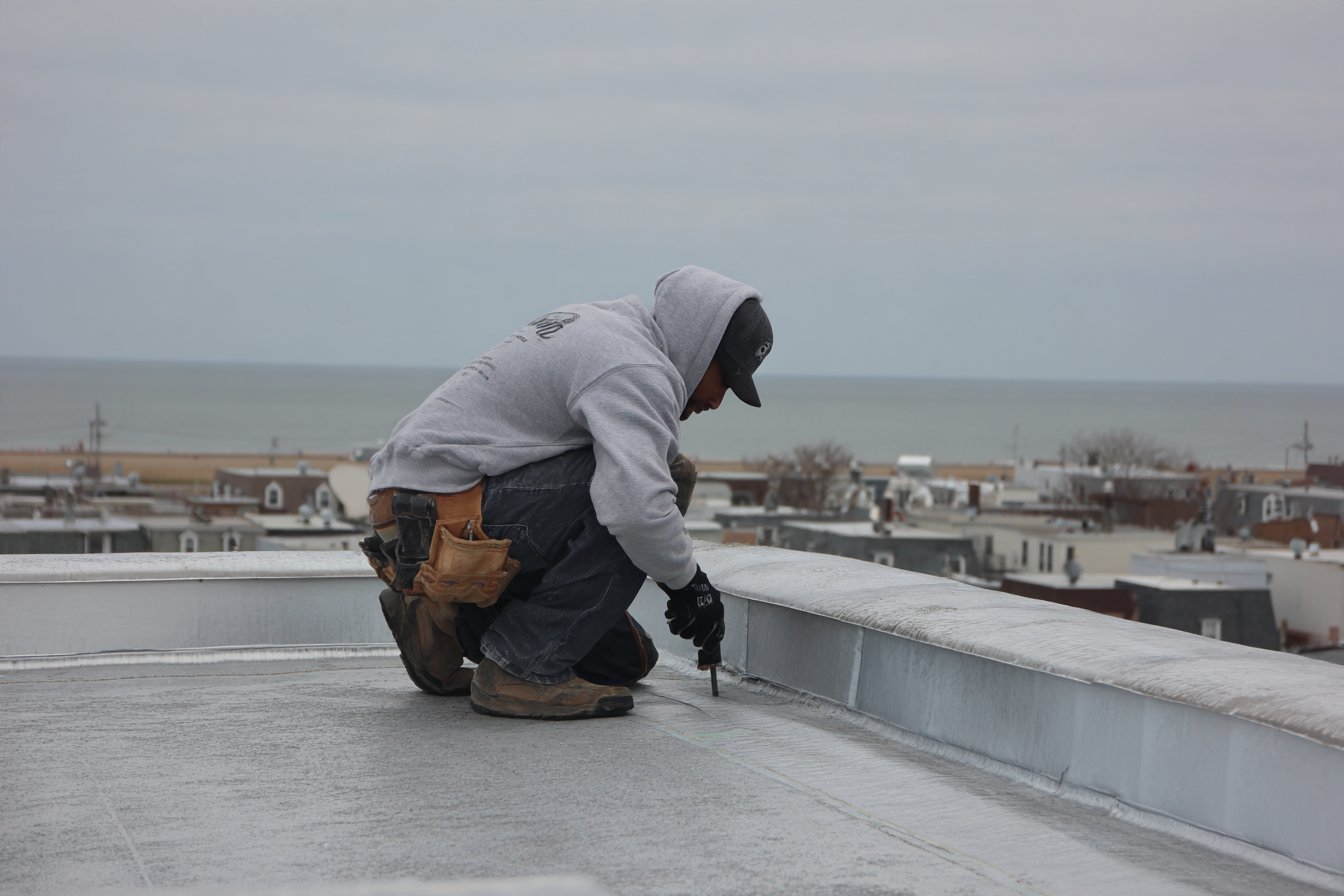 Roofer re-flashing a parapet on a flat modified bitumen roof in Far Rockaway, Queens, with the Atlantic Ocean beyond the rooftops