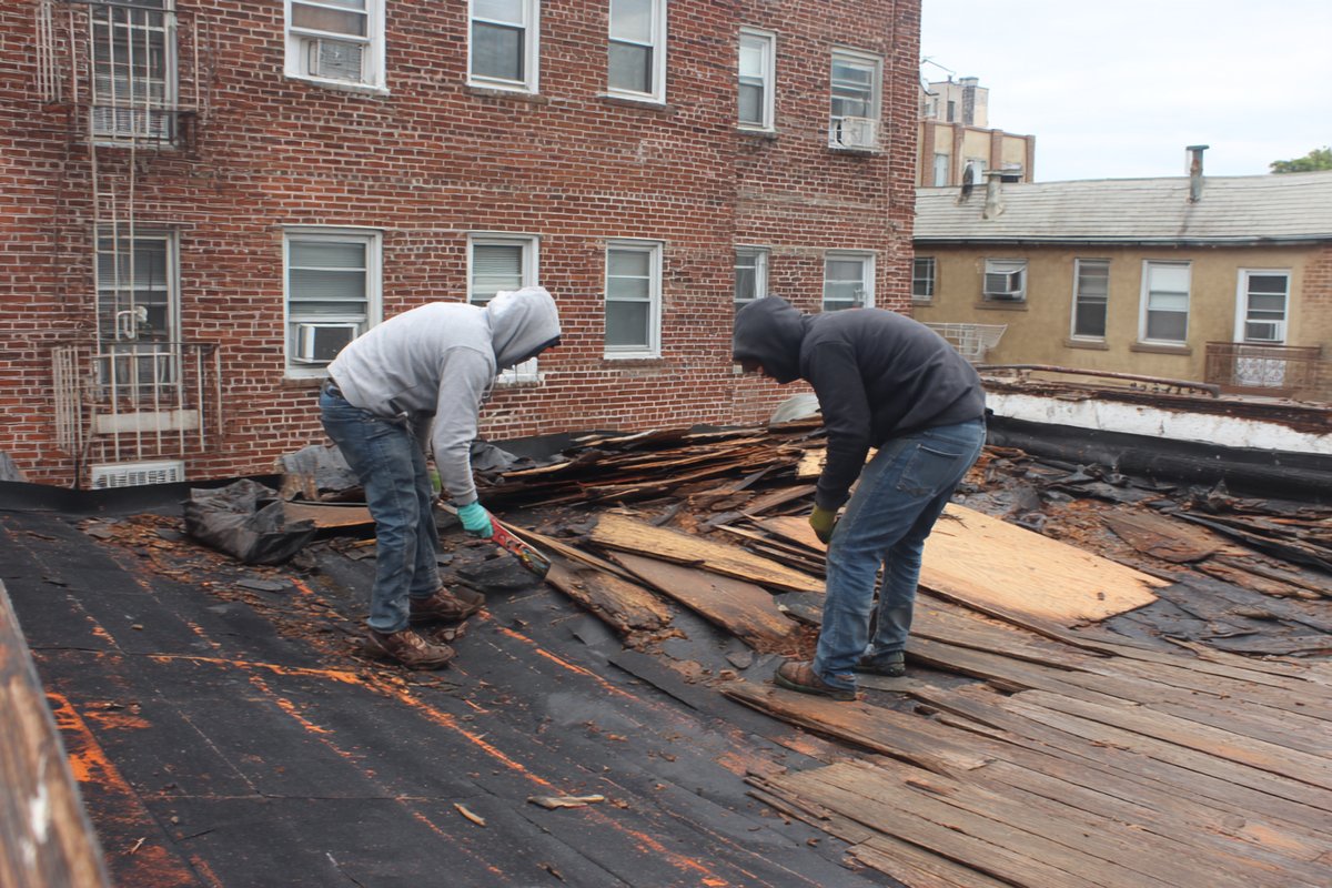 Roofers tearing an old flat roof down to the wood deck on a Queens brownstone, rotted boards marked for replacement