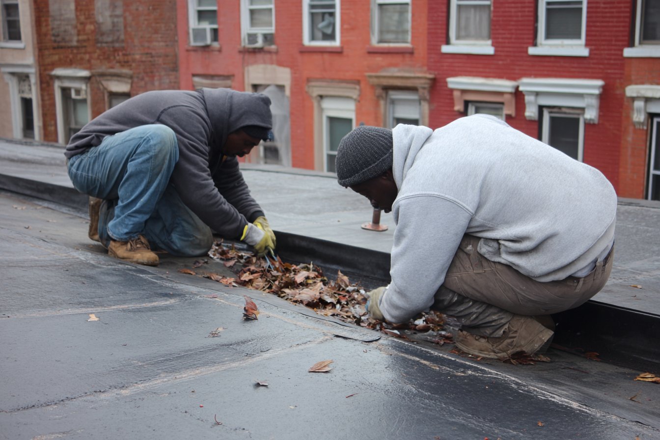 Flat Roofers NY crew clearing a roof drain and checking membrane seams during a spring maintenance visit on a Queens flat roof