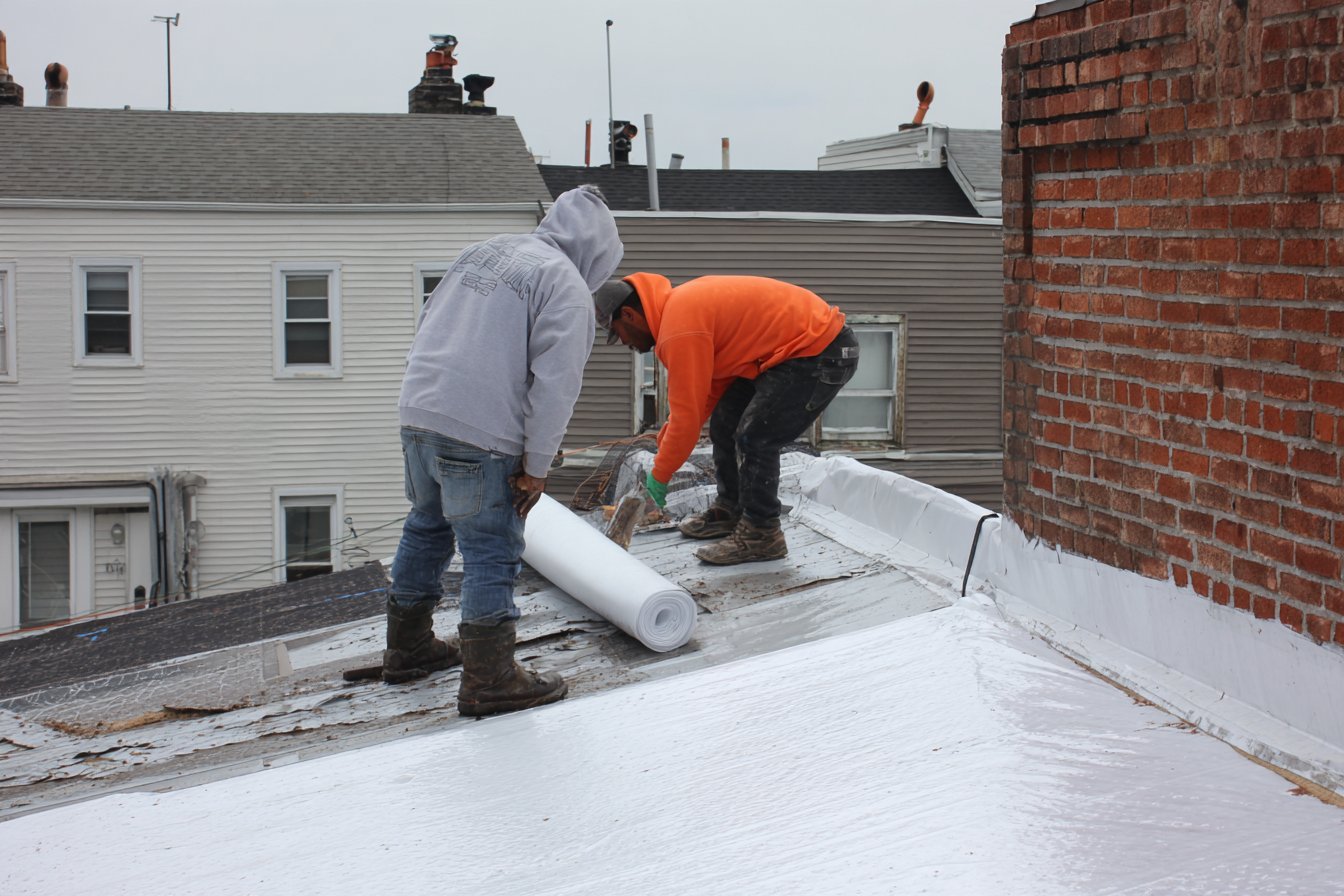 Crew rolling out a new white TPO membrane on a Queens row house roof after the old layers were torn off