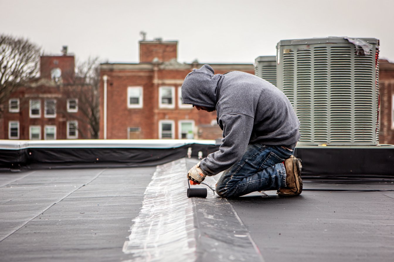 Flat Roofers NY roofer sealing a seam on the flat roof above a Queens storefront, rooftop HVAC unit behind him