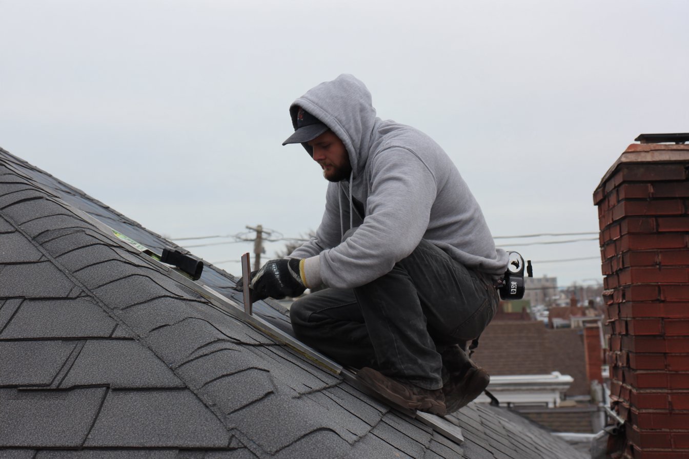 Roofer measuring the pitch of a sloped front roof above a flat main roof on a Queens rowhouse