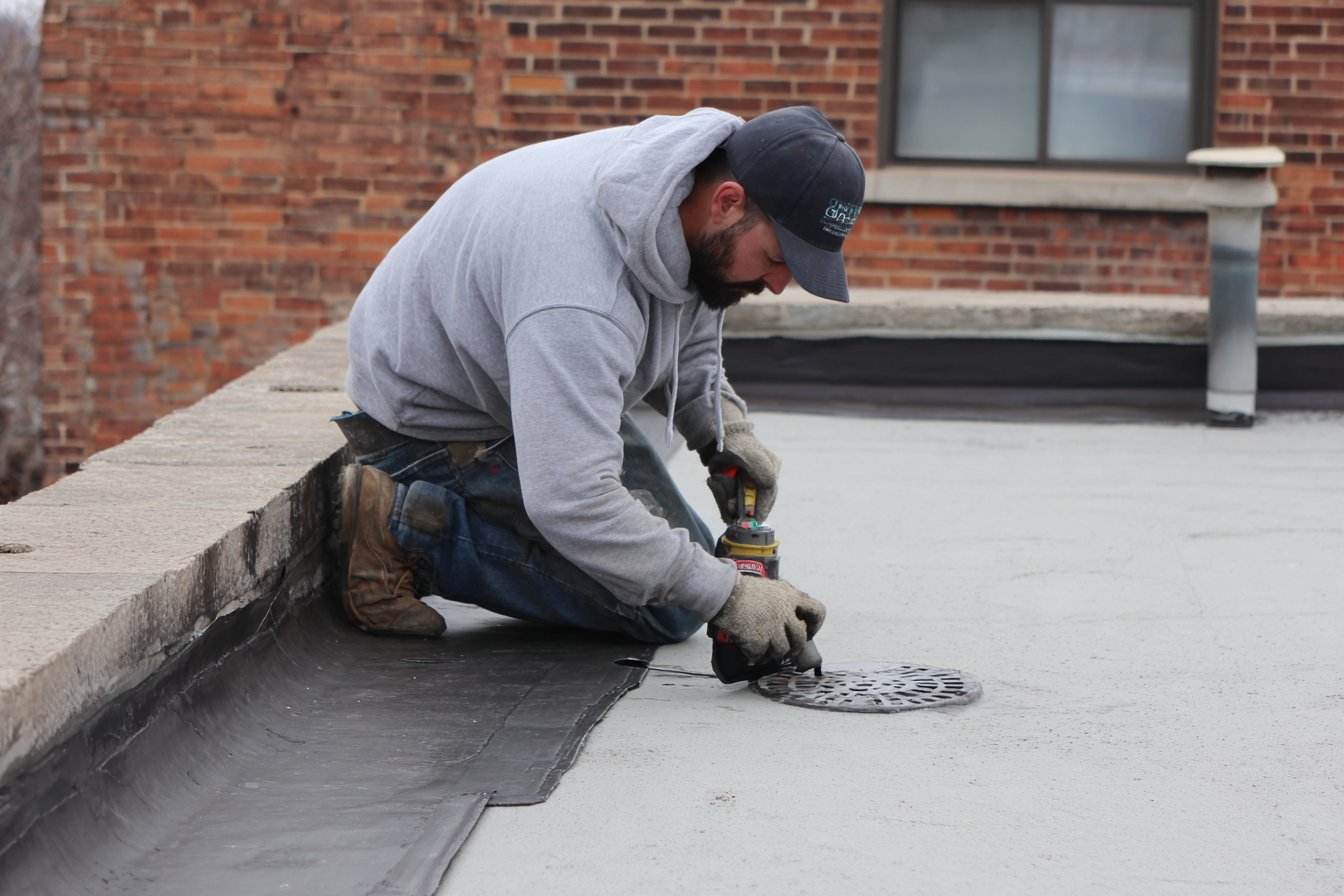 Flat Roofers NY roofer resealing an open seam on the flat roof of a Queens row house, parapet wall and roof drain in view