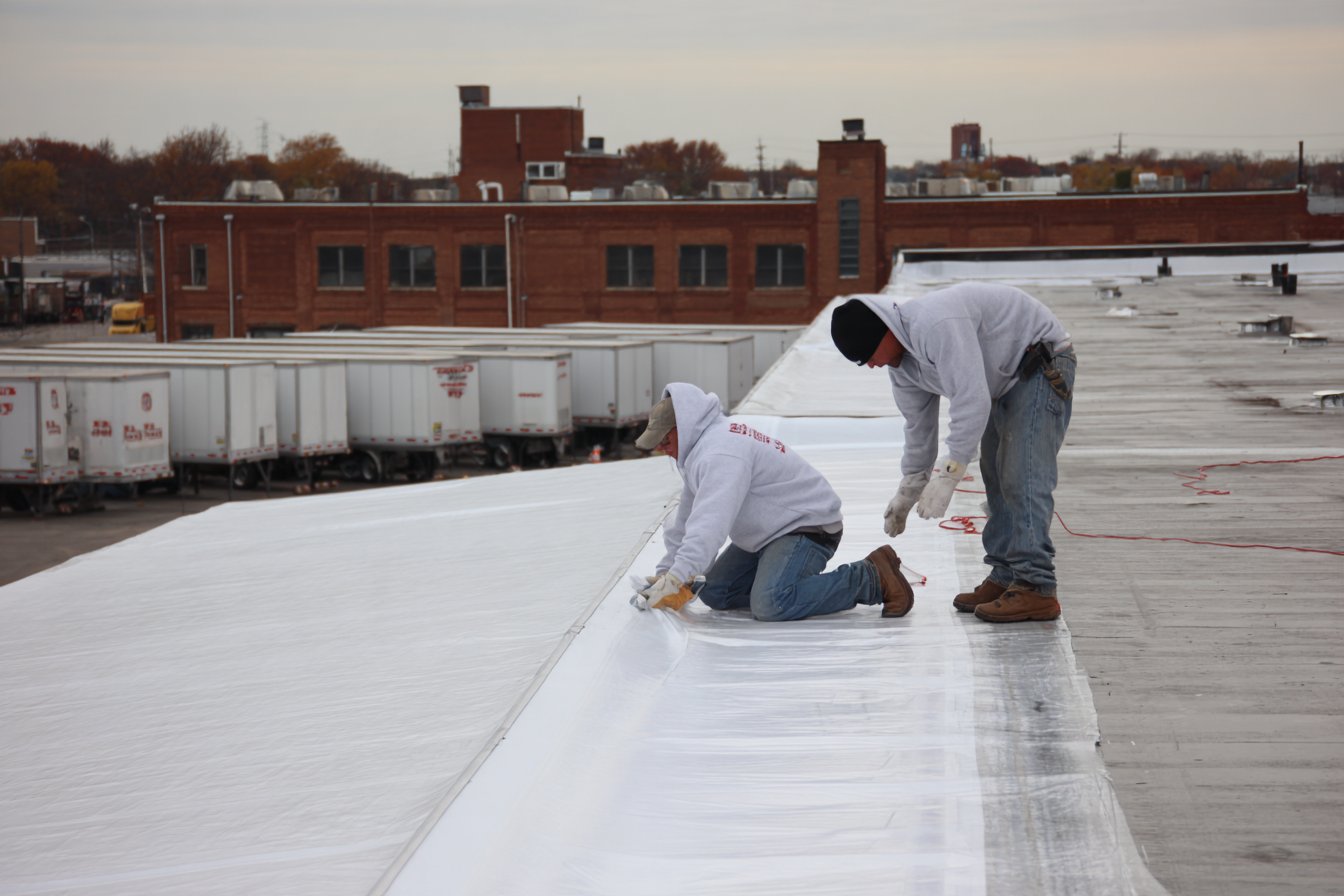 Flat Roofers NY crew installing a white TPO membrane on a large Queens warehouse roof while trucks load at the docks below