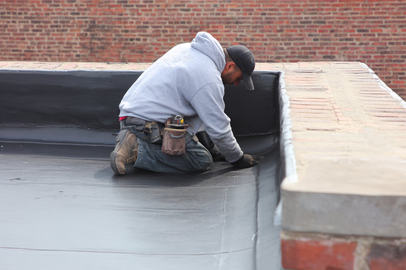Flat Roofers NY roofer on the flat modified bitumen roof of a converted brick warehouse loft in Long Island City, parapet wall in the foreground