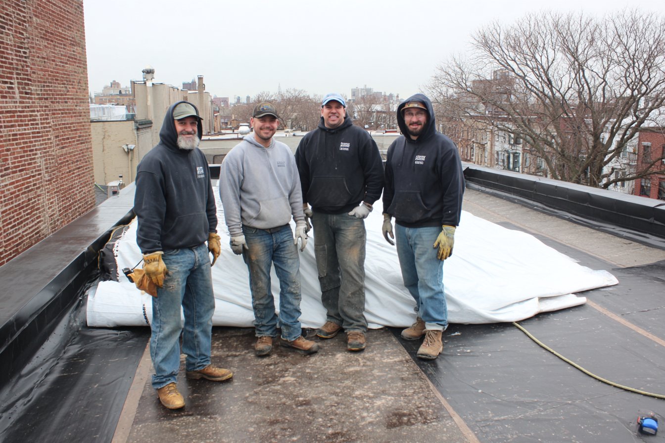 The Flat Roofers NY crew on a Queens flat roof, rolling out new membrane by the parapet