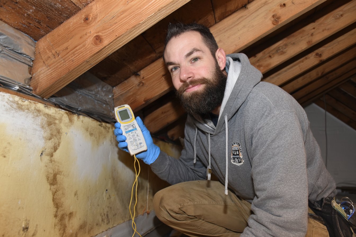 Roofer in the attic of a Queens house holding a moisture meter against water-stained roof sheathing under a sloped roof