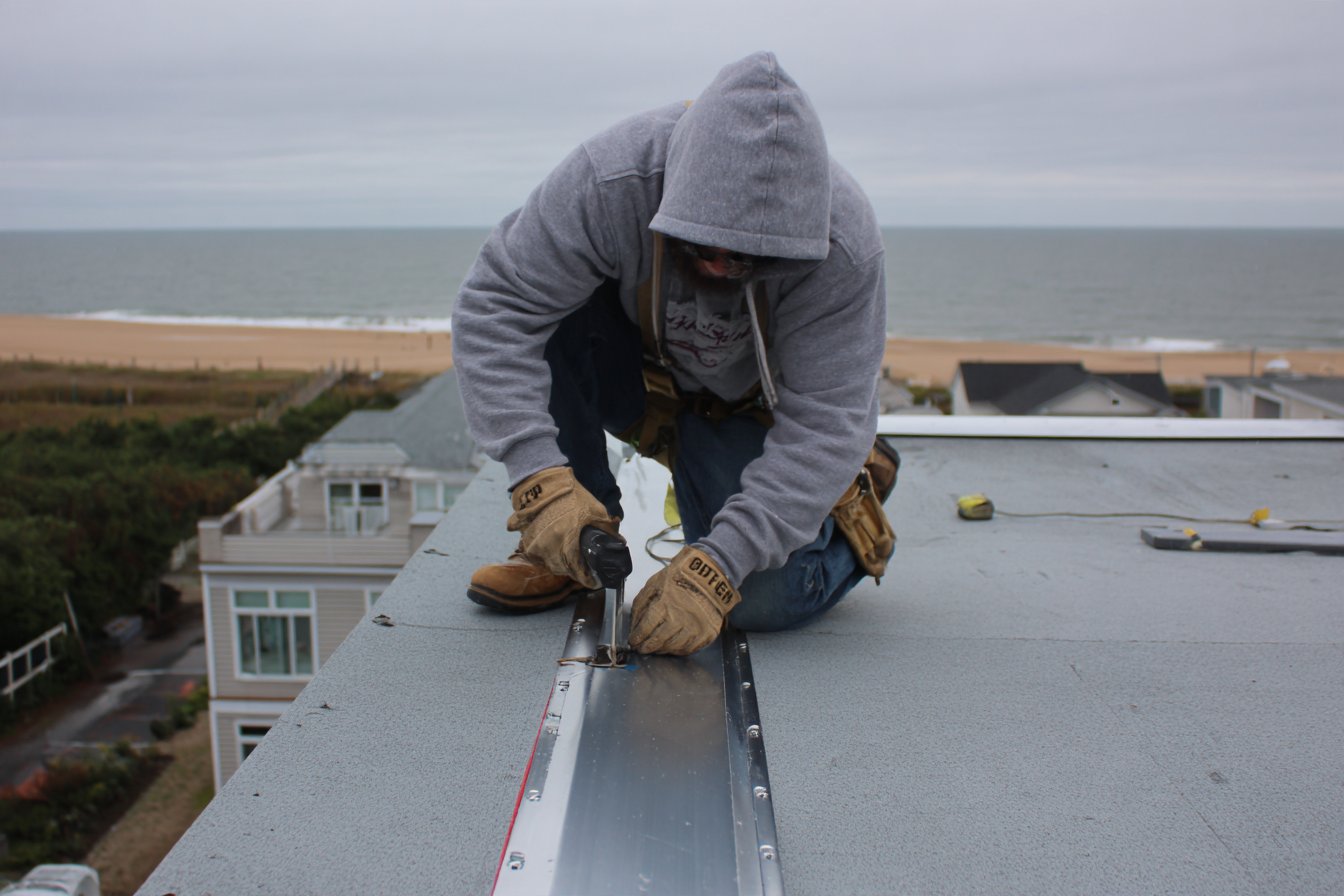 Roofer sealing the edge metal on a flat roof in Rockaway Beach, with the beach and the Atlantic Ocean a short block behind the house
