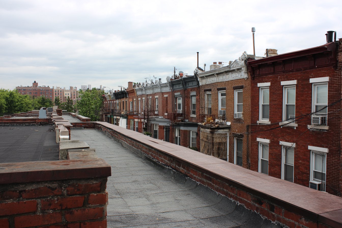 A row of attached Sunnyside brick rowhouses with joined flat roofs behind low parapet walls, one continuous roofline across the block