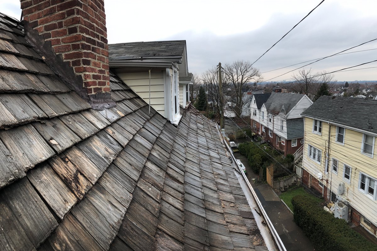 Weathered gray cedar shingle roof slope on an older Queens colonial, with a few freshly replaced boards blending in